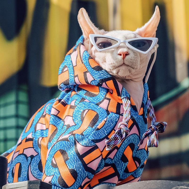 Sphynx cat wearing white cat-eye sunglasses and colorful patterned hoodie sitting outdoors
