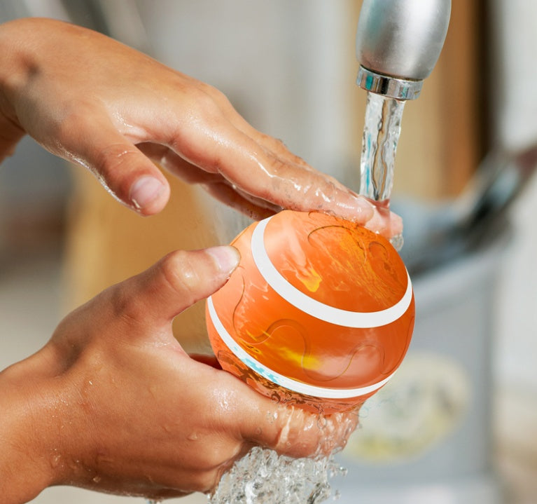 Hands washing an orange spherical puzzle toy under running water from a faucet